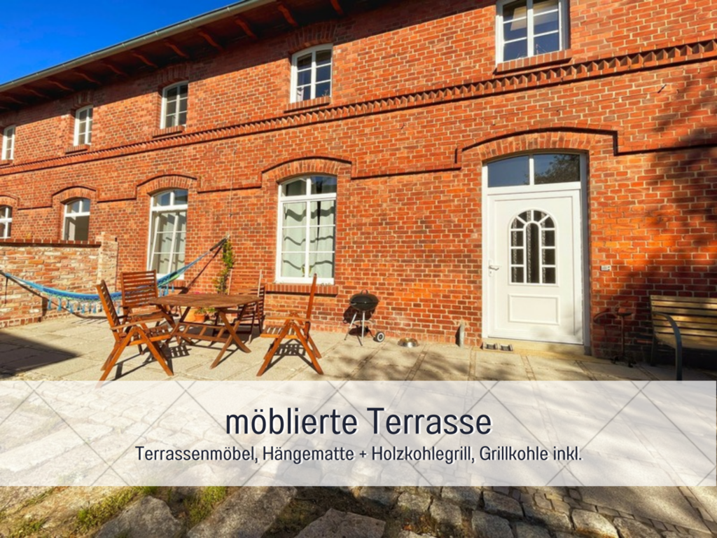 Eine möblierte Terrasse vor einem roten Backsteinhaus mit Holztisch, Stühlen, Hängematte und einem Holzkohlegrill.